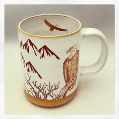 a coffee cup with an eagle painted on the side and mountains in the background, sitting on a white surface