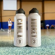 two white water bottles sitting on top of a basketball court next to each other in front of a woman