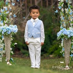 a little boy in a blue vest and white pants standing under an arch with flowers