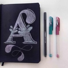 a black notebook with the letter a drawn on it next to two markers and pens