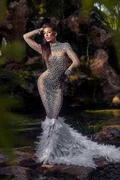 a beautiful woman in a long dress standing on rocks near water and palm trees with her hands behind her head