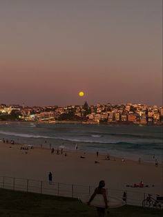 people are on the beach at sunset with buildings in the background and one person carrying a surfboard