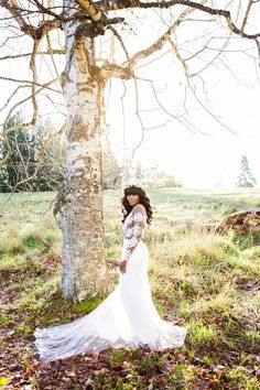 a woman standing next to a tree in a field with leaves on the ground and wearing a white dress