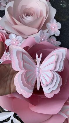 a hand holding a pink paper flower with a butterfly on the center and flowers in the background