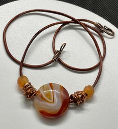 This vibrant necklace features a striking Sardonyx Agate pendant, known for its bold bands of color, complemented by warm copper and rich Carnelian beads. Strung on a durable leather cord, the natural textures and tones create a perfect balance of earthy elegance and bright, energetic design. Ideal for those who love statement pieces with a touch of artisanal flair, this necklace adds a pop of color and warmth to any outfit. Approximately 20" long. For reference Sardonyx Agate in almost 1" aroun Adjustable Carnelian Necklace Hand Wrapped, Adjustable Hand Wrapped Carnelian Necklace, Unique Adjustable Carnelian Necklace, Adjustable Brown Wire Wrapped Necklace, Adjustable Wire Wrapped Carnelian Necklace, Brown Agate Hand Wrapped Necklace, Adjustable Carnelian Round Pendant Necklace, Artisan Brown Necklace With Waxed Cord, Amber Adjustable Hand Wrapped Necklace