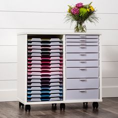 a white cabinet with drawers and flowers on top is shown in front of a wall