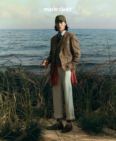 a man standing in front of the ocean wearing a hat and jacket with his hands in his pockets