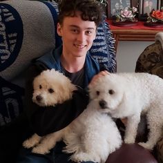 a young man sitting in a chair with two small white poodles on his lap