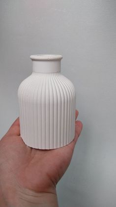 a hand holding a small white vase in front of a white wall with no one around it