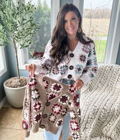 A woman smiling, holding a knitted sweater with granny square patterns, standing indoors beside a window and a potted plant. Grant Square Cardigan, Granny Square Jumper Pattern Free, Granny Square Crochet Sweater Pattern, Crochet Granny Square Sweater Pattern, Granny Square Sweater Pattern Free, Granny Square Projects Ideas, Granny Square Sweater Pattern, Granny Square Crochet Sweater, Crochet Cardigan Free Pattern