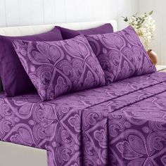 a bed with purple bedspreads and pillows on it in a white room next to a vase