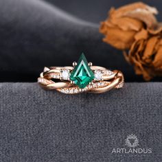 a ring with an emerald colored stone surrounded by white diamonds on a gray surface next to a dried flower