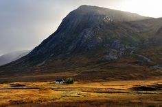 a house in the middle of an open field with mountains in the backgroud