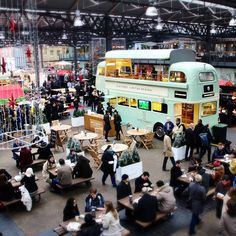 a crowd of people standing around tables and benches in front of a double decker bus