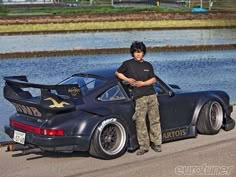 a man standing next to a black sports car on the side of a road near a body of water