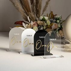 three clear acrylic signs with gold writing on them and flowers in the background