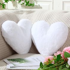 two white heart shaped pillows sitting on top of a couch next to flowers and a book