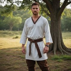 a man dressed in white and brown standing next to a tree with his hands on his hips