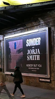 two people walking down the sidewalk in front of a movie poster on a building wall