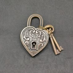 a heart shaped lock with two keys attached to it on a gray surface, the key is in the shape of a lion's head