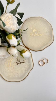 two wedding rings and some flowers on a white surface with the word vera written in gold