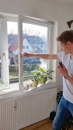 a young man standing in front of a window looking at his cell phone and pointing to the outside