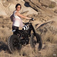 a woman standing next to a dirt bike in the middle of a field with dry grass