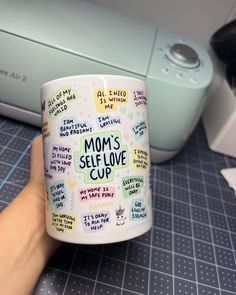 a person holding up a coffee mug with words on it and the word mom's self love cup