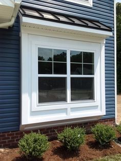 a blue house with white trim and windows in the front yard, shrubs around the window