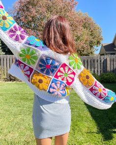 a woman standing in the grass with her back to the camera, wearing a colorful crocheted sweater