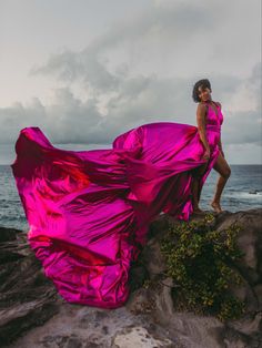 a woman in a pink dress standing on rocks near the ocean with her flowing fabric