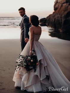 a bride and groom standing on the beach at sunset with their wedding dress blowing in the wind