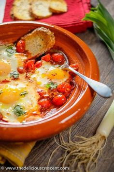 an orange bowl filled with eggs and tomatoes on top of a table next to bread