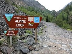 a sign that is on the side of a dirt road with rocks and trees in the background