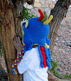 a little boy wearing a crocheted blue hat with red and yellow horns standing next to a tree