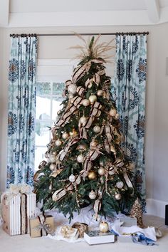 a decorated christmas tree in front of a window with blue drapes and gold ornaments