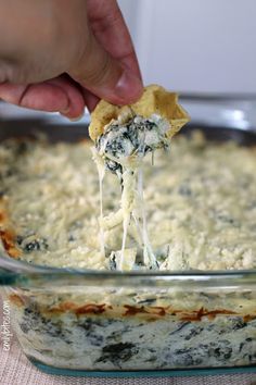 a hand holding a tortilla chip over a casserole filled with spinach and cheese