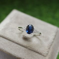 an oval blue sapphire and diamond ring sits on a white velvet box with grass in the background