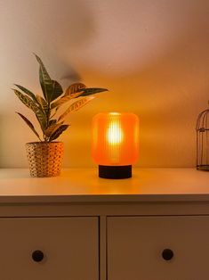 an orange lamp sitting on top of a white dresser next to a potted plant