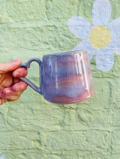 a person holding a purple coffee mug in front of a green brick wall with flowers painted on it