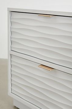 a white chest of drawers with gold handles and wavy lines on the front, in an empty room