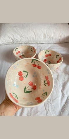 three bowls with cherries painted on them sitting on a bed next to a person's hand