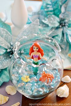 a glass bowl filled with little mermaids on top of a table next to shells