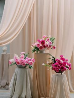 two vases filled with pink flowers sitting on top of a table next to drapes