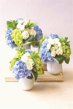 three white vases filled with blue and green flowers on a wooden tray next to each other