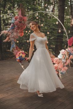 a woman in a white dress is standing on a wooden deck with flowers and an arch