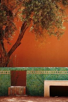 a bench under a tree in front of a red wall with green tiles on it