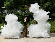 a woman standing next to two large white flowers on top of a cement ground with trees in the background