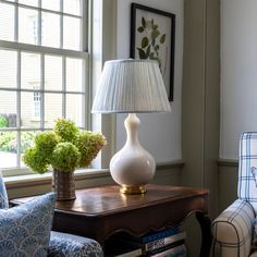 a lamp sitting on top of a wooden table next to a chair and ottoman in front of a window