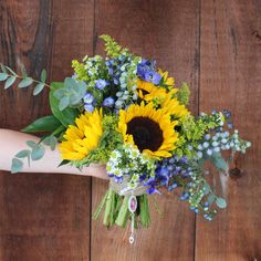 a bouquet of sunflowers and blue flowers on a wooden table with greenery